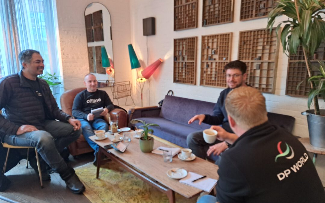 Vier junge Männer sitzen in einer Vintage Hotel-Lobby am Tisch - der Mann mit dem Rücken zum Fotografen hat das Logo DP World auf der Jacke