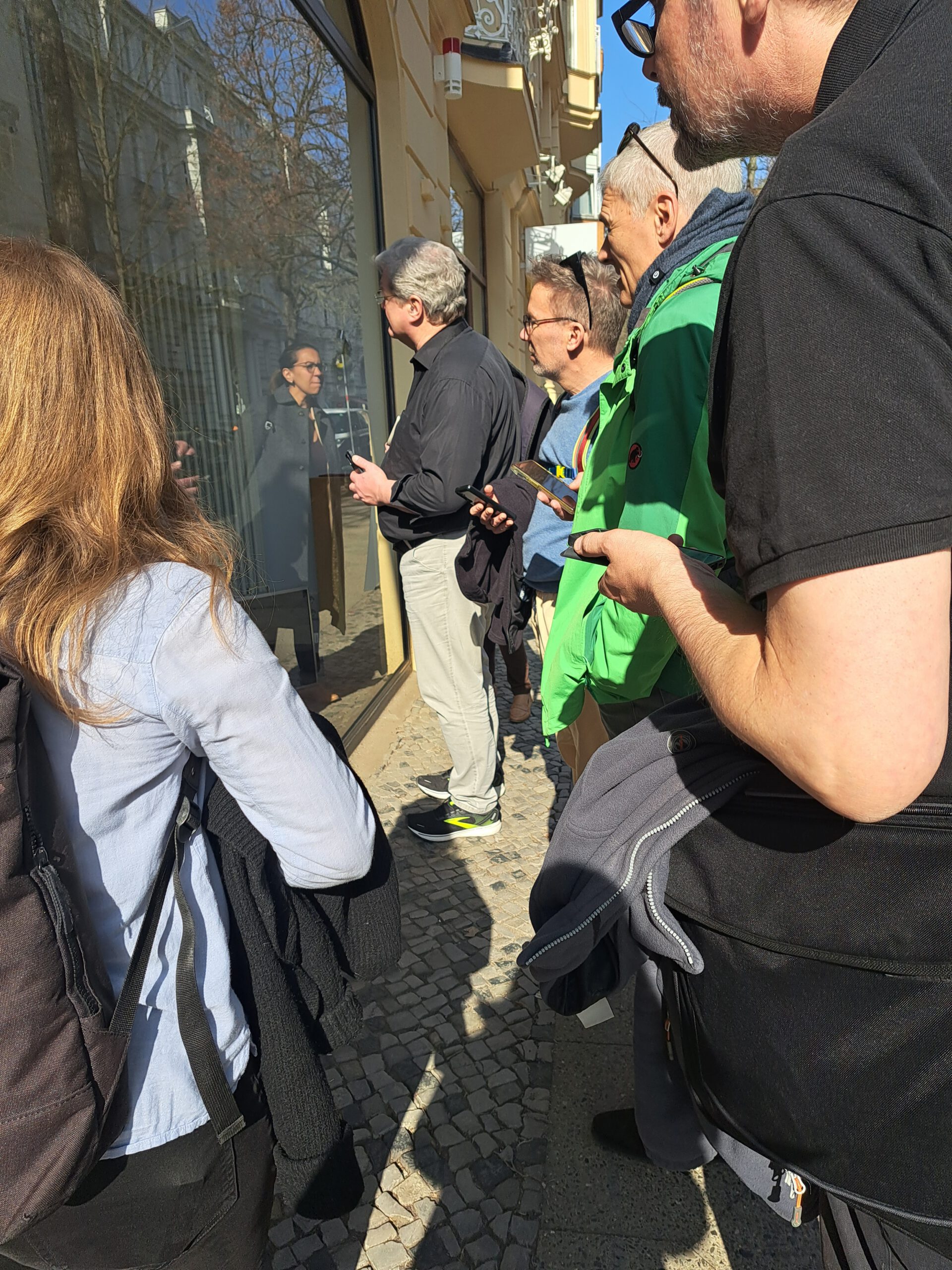 vier grauhaarige Männer blicken konzentriert durch Fensterscheibe während im Vordergrund mit dem Rücken zum Betrachter eine Frau steht und eine weitere Frau ist nur als Spiegelbild im Fenster erkennbar